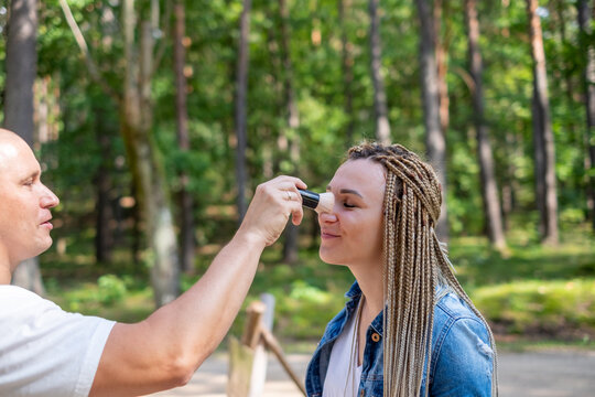 A man applies make up with a brush to a woman's nose in a wooded area, depicting care and helping. Suitable for health and wellness, skincare, and outdoor activity themes.