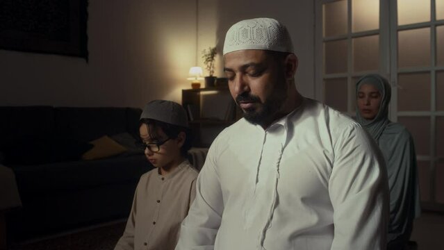 Side footage of Biracial man in muslim cap and thawb praying to Allah together with his son and wife who sitting behind them during namaz at home
