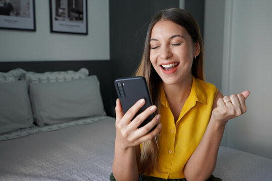 Young Brazilian woman excited for success holding smartphone with fist up and eyes closed celebrating victory smiling in her room at home. Winner concept.
