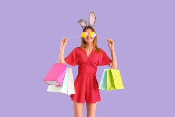 Young woman in bunny ears with shopping bags and paper Easter eggs on lilac background