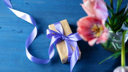 A gift box in craft paper with a ribbon on a blue table. Pink tulips in a vase. Top view, copy space