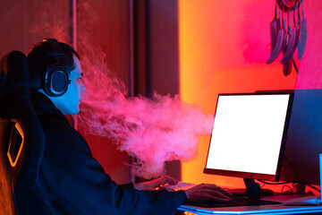 A young man wearing headphones sits at a computer and plays games. Vapes and exhales a cloud of...