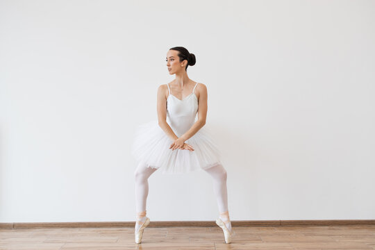 Beautiful young female dancer posing on studio background. Caucasian ballerina in white bodysuit and tutu poses in motion showing ballet elements while standing on pointe shoes.