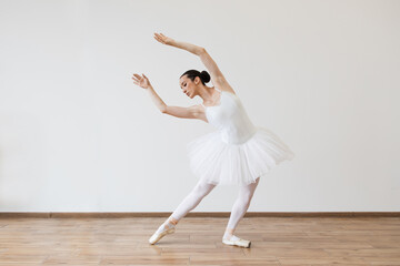 Caucasian young woman ballerina in white tutu, dancing on pointe with arms overhead, in the studio against a light bright background of modern studio hall.