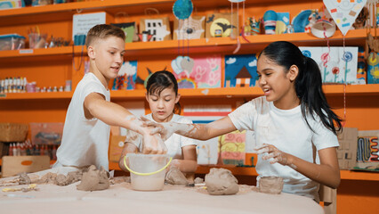 Handsome student dipping hand in to water to soften clay at art classroom. Group of diverse children working or modeling vase. Happy boy put water to young girl while laugh with happy. Edification.
