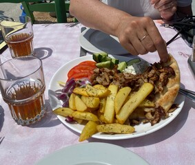 Plate of souvlaki, fries, pita, and salad on a table with drinks, hand takes from the pita to eat