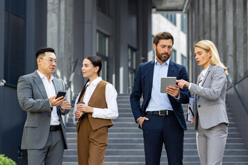 A group of four professionals engaging in a discussion outside a contemporary office building, with digital tablets and smartphones in hand, embodying teamwork and collaboration.