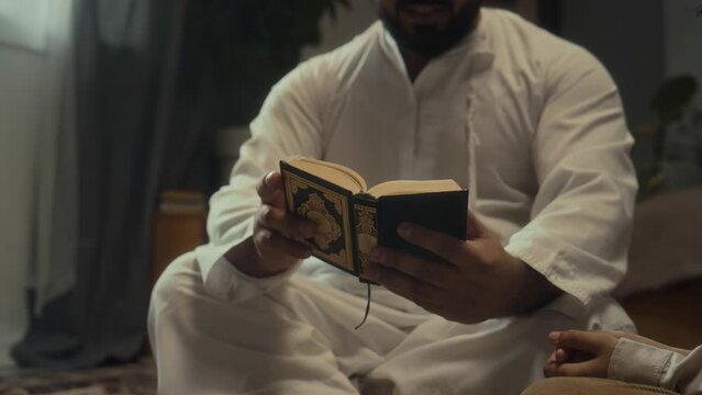 Cropped footage of unrecognizable bearded religious man sitting on floor and paging through mini Al Quran while spending evening with family
