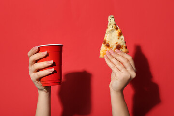 Female hands holding tasty pizza slice and one-use cup on red background