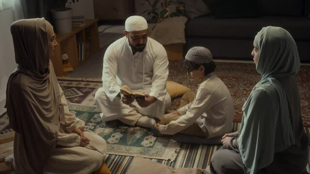 High angle footage of muslim family members sitting on floor and listening to head of family reading prayers and basic rules from mini Koran and educating little son at home