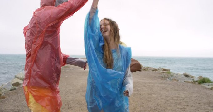 People, dancing in rain and beach celebration for holiday, winter vacation and party, freedom or energy. Young group of friends with raincoat and carefree, fun or excited for adventure by ocean