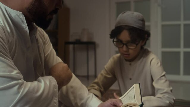 Side tilt footage of Biracial bearded father in white kurta reading Koran and explaining its rules and principles to little son who constantly nodding