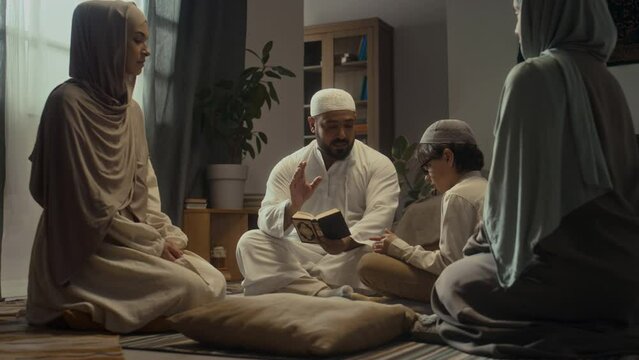 Ground level handheld footage of covered daughter and mother watching and listening how father in white thobe giving religious education to little son explaining Quran rules at home