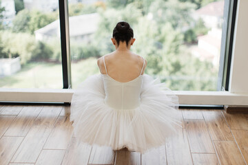 Fototapeta premium Classical Ballet dancer. Beautiful Caucasian graceful female ballerina in tutu skirt sitting at wooden floor and stretching while practice split ballet position, back view.