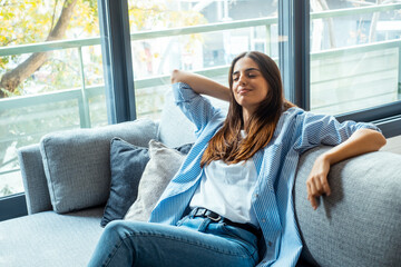 Kick back and relax concept. Young beautiful brunette woman with blissful facial expression alone on the couch with her bare feet on coffee table. Portrait of relaxed female resting at home. - Powered by Adobe