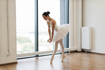 Young Caucasian classical ballet dancer woman in dance class. Beautiful graceful ballerina practice ballet positions in white tutu skirt near large window in white light hall.