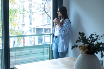 One woman at home standing and looking outside the window drinking cup of coffee and smiling. One female young adult people living alone. Millennial thinking and relax indoor in modern apartment town