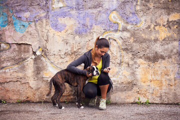 young woman playfully interacting with a Boxer dog, against a graffiti-covered wall in an urban setting. - 750972545