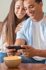 Smiling young couple embracing while looking at smartphone. Multiethnic couple sharing social media on smart phone. Smiling african girl embracing from behind her happy boyfriend while using cellphone