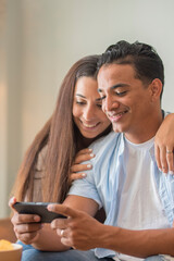 Smiling millennial couple sit on sofa in living room have fun using modern smartphone devices together, happy young husband and wife laugh relax at home browsing application on cellphone gadgets web