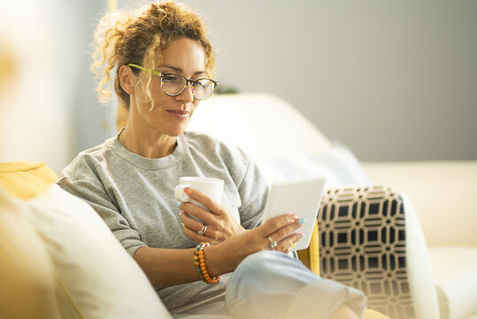 Smiling middle-aged Caucasian woman sit on couch in living room browsing wireless Internet on tablet, happy modern pretty female relax on sofa at home using pad device, yellow, technology concept