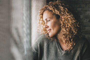 One serene adult woman looking outside the window at home with peaceful expression on face and little smile. Confident female people in indoor relax leisure activity alone. Dreaming and thinking lady - Powered by Adobe