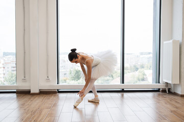 Caucasian ballerina stretching to feet while standing in dance studio with large windows. Young...