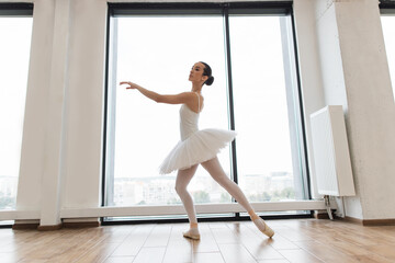 Fototapeta na wymiar Beautiful ballerina practice ballet positions in white skirt near large window in white hall. Young ballet dancer - Harmonious pretty woman with tutu posing in studio - Contemporary dance performer.