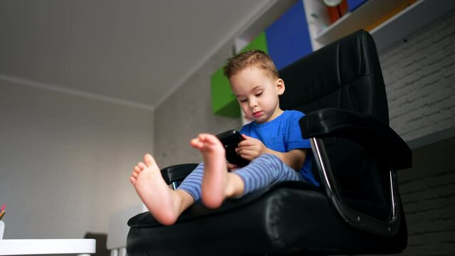 Small kid sitting in the big chair looks at smartphone. Low angle view on the barefoot toddler focused on the gadget.