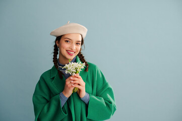 Fashionable happy smiling woman wearing white beret, trendy green coat, holding spring bouquet of snowdrops, posing on blue background. Studio portrait. Copy, empty, blank space for text - 750980118