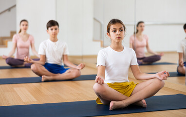 Portrait of focused teen girl meditating in yoga position Padmasana during family training at gym..