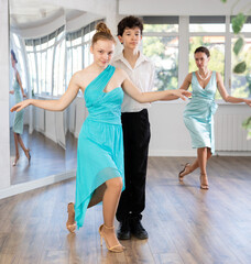 Couple of successful elegantly dressed young dancers, teenage girl and boy performing curtsy and bow after dance performance in choreography studio with female teacher in background..