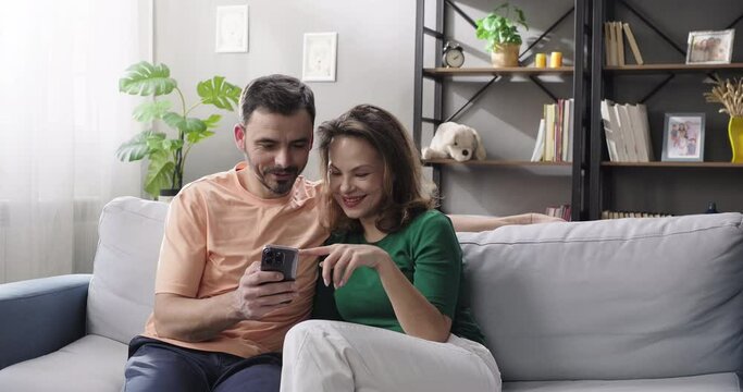 Happy couple using smartphone, shopping online, ordering delivery, planning vacation, booking hotel, checking social media apps, watching videos while sitting on couch in the living room