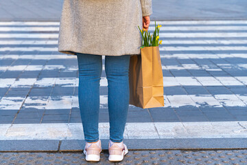 Girl woman stands at a pedestrian crossing and holds a paper bag with yellow daffodil flowers in her hands.She walks around the city center in the spring.