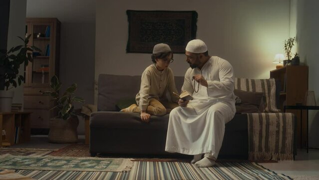 Full footage of muslim father in white kandora reading prayers and rules from Quran and holding tasbih while educating his little son to use beads in living room in evening