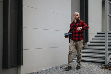 A focused entrepreneur outdoors checks his watch as he carries a notebook, seemingly late for a...