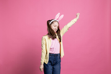 Positive enthusiastic girl fooling around and dancing on camera, wearing bunny ears in studio and showing dance moves. Young toddler acting cheerful and excited about easter celebration.