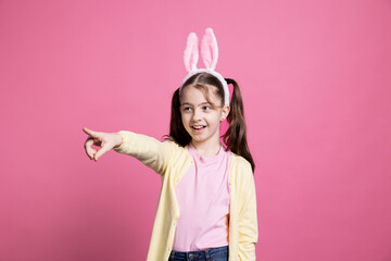 Cheerful young kid with pigtails fooling around in front of camera, pointing at something in studio...