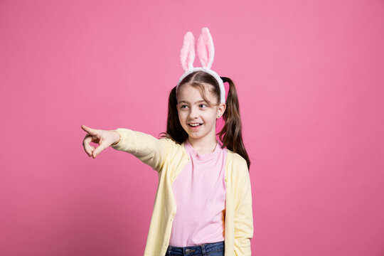 Cheerful Young Kid With Pigtails Fooling Around In Front Of Camera, Pointing At Something In Studio And Dancing. Positive Excited Schoolgirl Feeling Confident While She Is Wearing Bunny Ears.