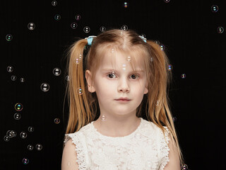 Child blonde girl 6 years old in a white dress on a dark background among air bubbles, photo studio,