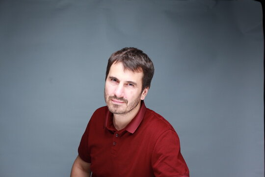 A picture of a man with a beard, mustache and fashionable hairstyle, dressed in a casual polo shirt, posing in a studio with a serious expression on a light background