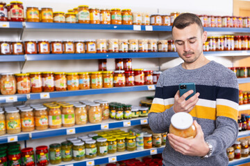 Man in store of imported Russian goods chooses jar with canned fermented cabbage. Male buyer photographs item, compares appearance of product in store and in mobile application