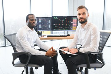 Two diverse colleagues traders talking to each other, sitting in the office in front of multiple computer screens. Stock trading, people, business concept