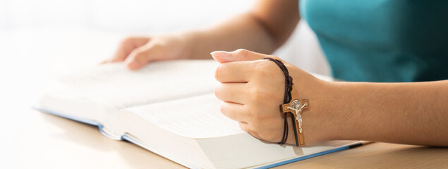 Cropped image of female reading a bible book while holding cross at wooden table with blurring...