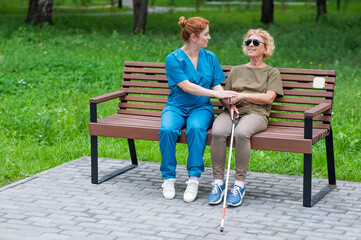 A nurse and an elderly blind woman are sitting on a bench in the park.