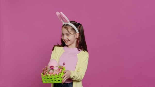 Smiling pleased girl presenting her handcrafted easter basket filled with painted eggs and other festive decorations for holiday celebration. Joyful small kid proud of her arrangement. Camera A.