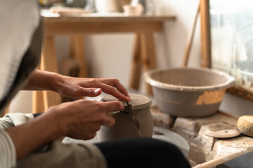 A close-up of a ceramist's hands as she meticulously trims a clay piece on a pottery wheel,...