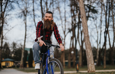 Bearded hipster man in a plaid shirt riding a bicycle in a tranquil urban park, embodying active lifestyle and leisure.