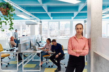 Fototapeta na wymiar Portrait of young smiling business woman in creative open space coworking startup office. Successful businesswoman standing in office with copyspace. Coworkers working in background