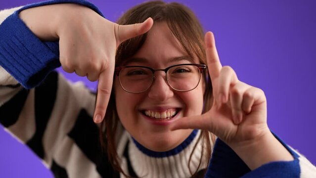 Closeup portrait of attractive happy young woman 20s smiling holding up hands as if framing a composition for camera or video isolated on purple background